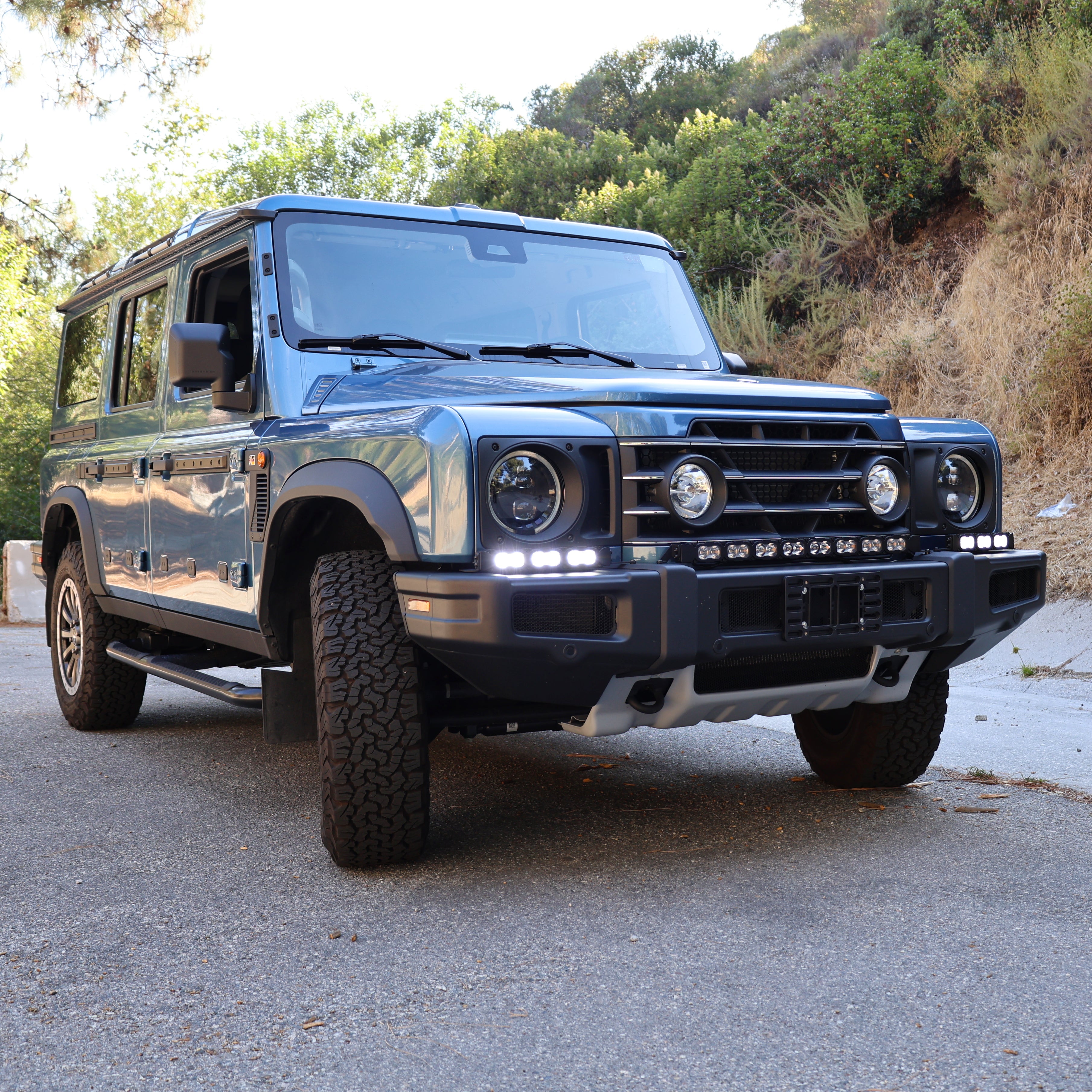 Blue 2024+ Ineos Grenadier front three-quarter view with XL light bar kit lit white on the front bumper