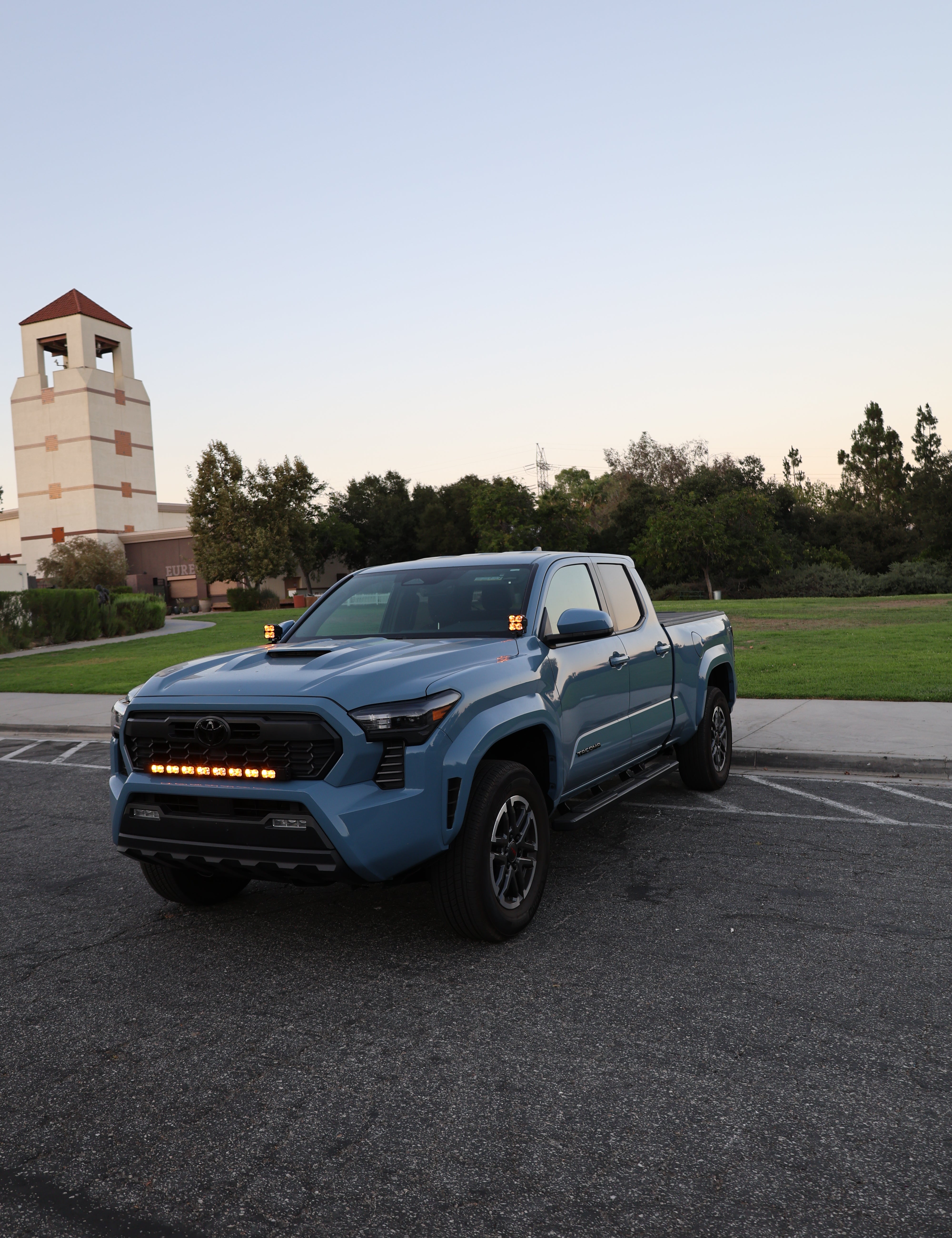 Light blue 2024+ Toyota Tacoma parked in a lot at dusk with amber DRL ditch lights glowing at the windshield cowl and matching amber light bar in the grille