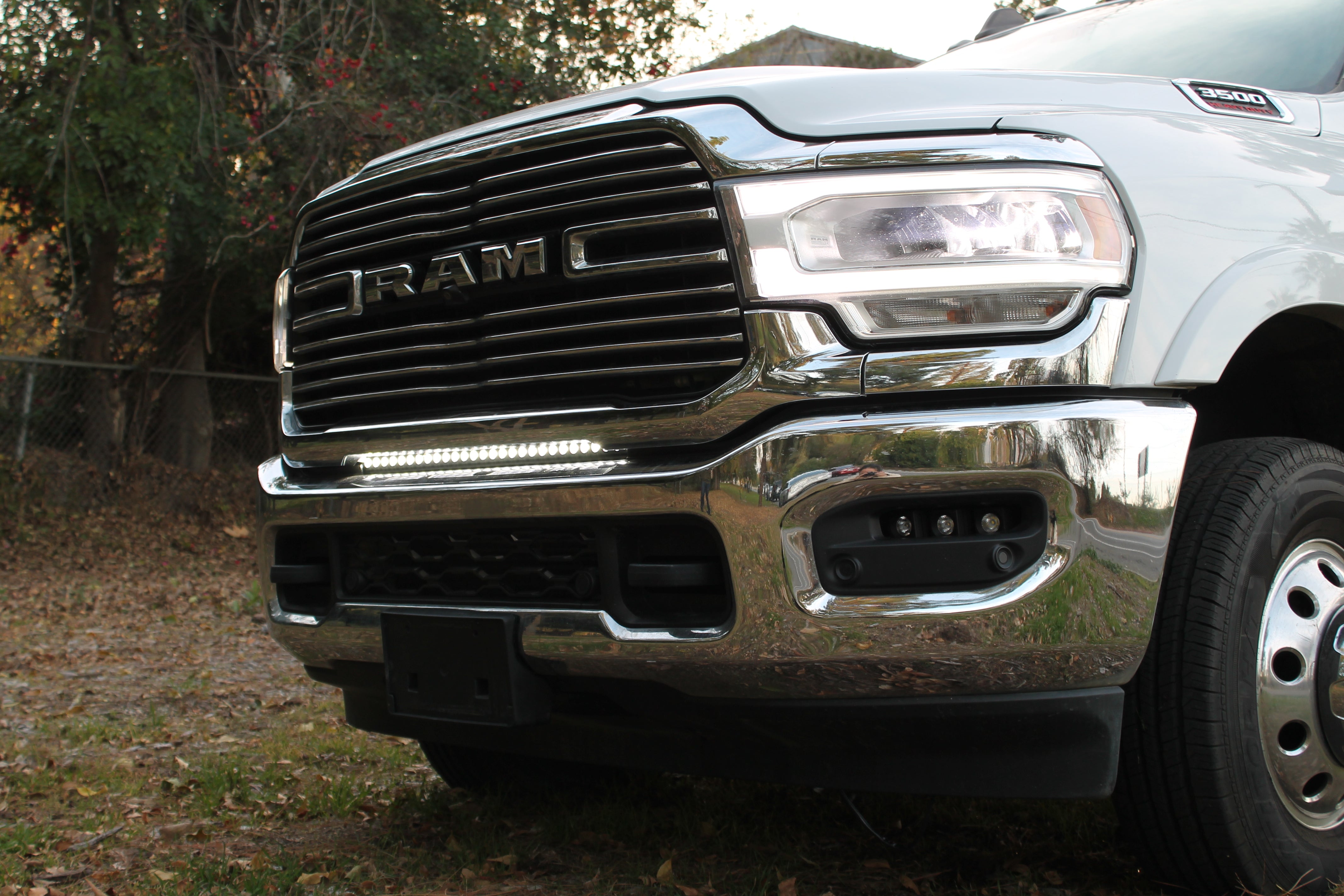 Close-up of the 30in light bar lit white below the chrome grille of a white 2019-2024 RAM 3500 dually, showing bracket fitment