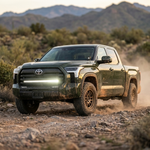 Dark green Toyota Tundra crew cab in action on a rugged rocky desert trail, front three-quarter view, featuring a bright white LED light bar mounted across the lower grille. Mud-splattered lower body, bronze wheels and all-terrain tires, additional ditch lights on the A-pillars, blurred mountains and scrub brush in the background under soft daylight.