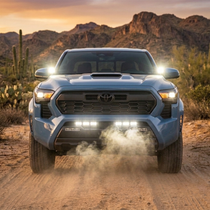 Light blue 2024+ Toyota Tacoma with dual 12in white LED light bars illuminated at the front bumper grille, driving on a desert dirt trail with mountains at sunset