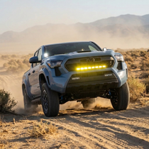 Light blue 2024+ Toyota Tacoma catching air off-road with a yellow LED 30in light bar illuminated above the grille, kicking up dust on a desert trail