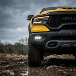 Yellow 2019-2024 RAM 1500 TRX on muddy off-road terrain with three white LED fog light pods lit in the front bumper, low front corner view