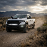 White 2025+ RAM 1500 Rebel driving a desert dirt road with the fog light kit's white LED pods lit in the front bumper, front three-quarter view
