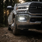 White 2019-2024 RAM 2500 on a wooded trail at dusk with three white LED fog light pods lit in the chrome front bumper, front corner view