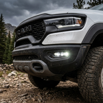 Close-up of the 2025+ RAM 1500 Rebel fog light kit with three white LED pods lit in the passenger-side front bumper of a mud-splattered white Rebel on a rocky trail