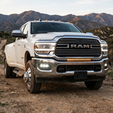 White 2019-2024 RAM 3500 dually on a mountain dirt road with white LED fog light pods lit in the chrome front bumper, front three-quarter view