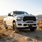 2025+ RAM 2500 lower grille pods with amber DRL lit, white truck on a desert trail in daylight, front three-quarter view